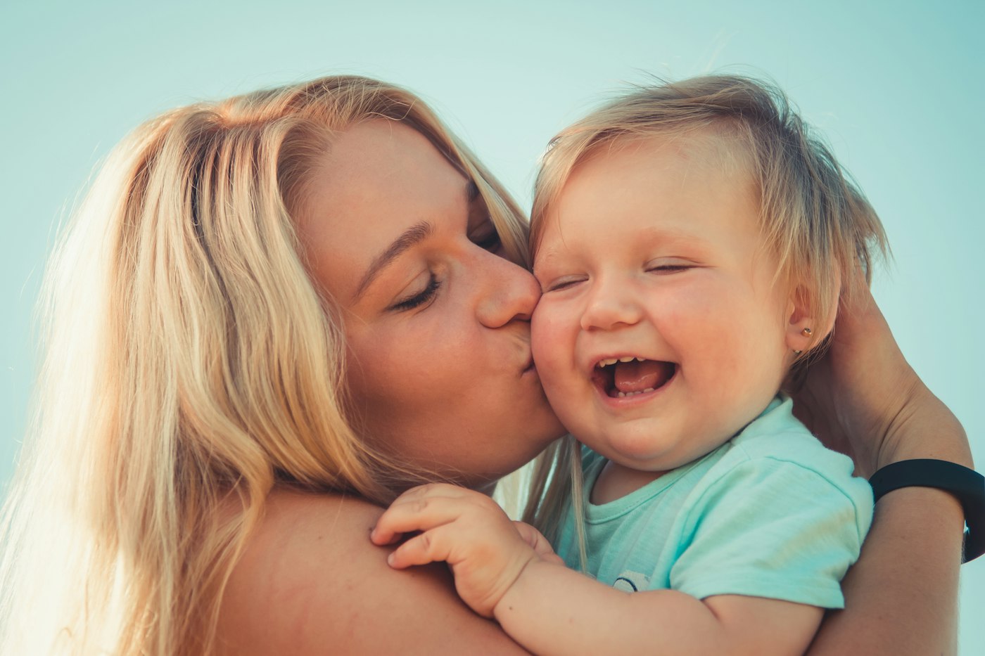 Mãe beijando a bochecha do filho bebê, que ri de olhos fechados