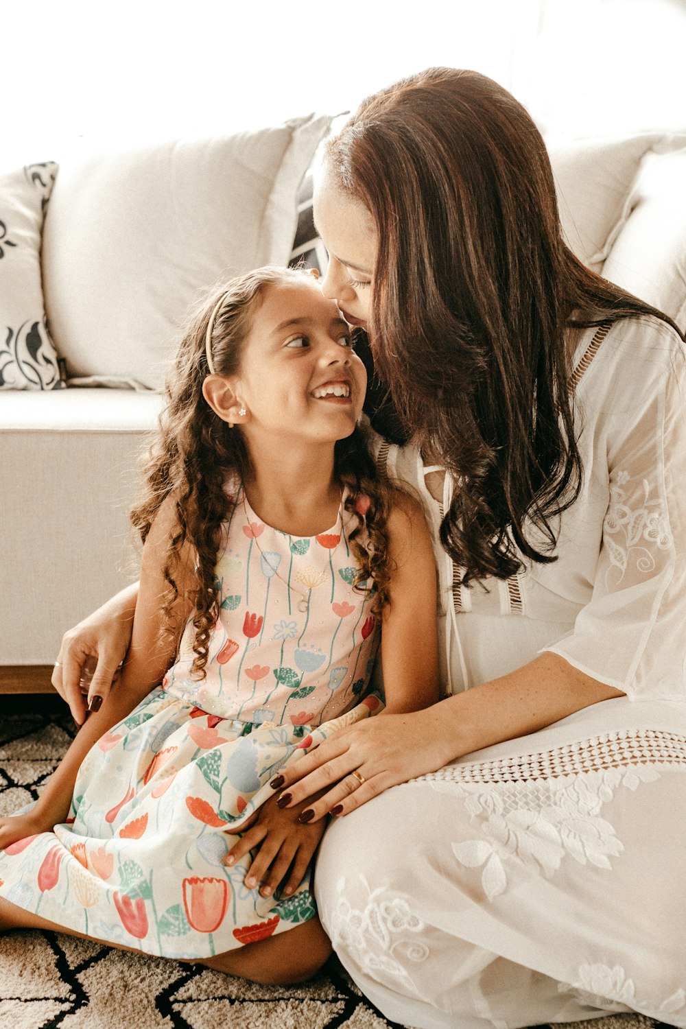 Mãe beijando a cabeça da filha, as duas sentadas num tapete